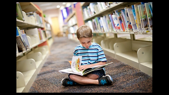 kid reading book on floor in library between rows of library shelves